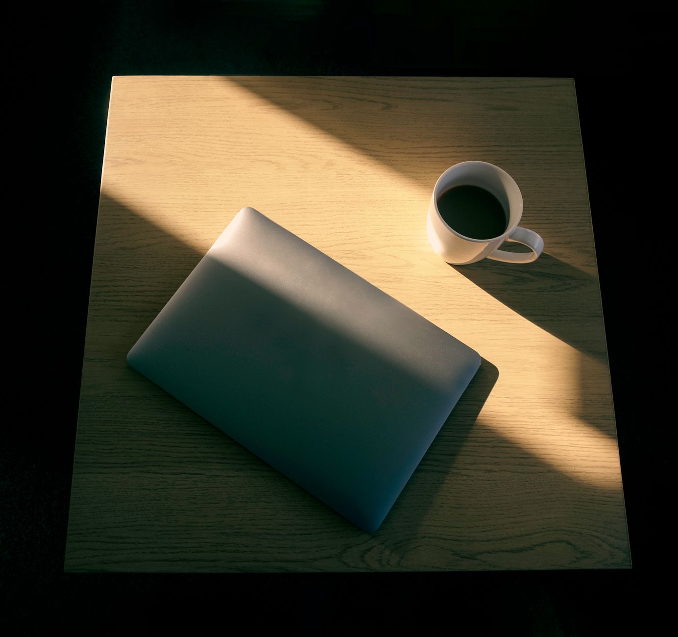 En lukket bærbar computer og en kop kaffe i skarpt sollys på et bord